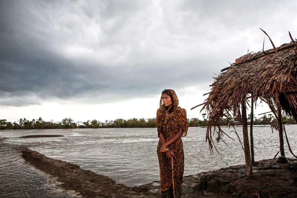 A young woman stands beside a thatched roof shack and looks out over flooded land