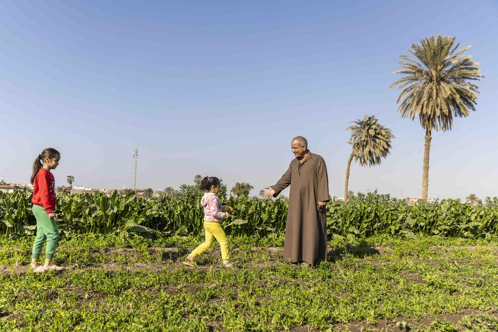 A man dressed in a brown tunic holds his hand out to two young girls who walk towards him. Two palm trees and green vegetation can be seen behind them.